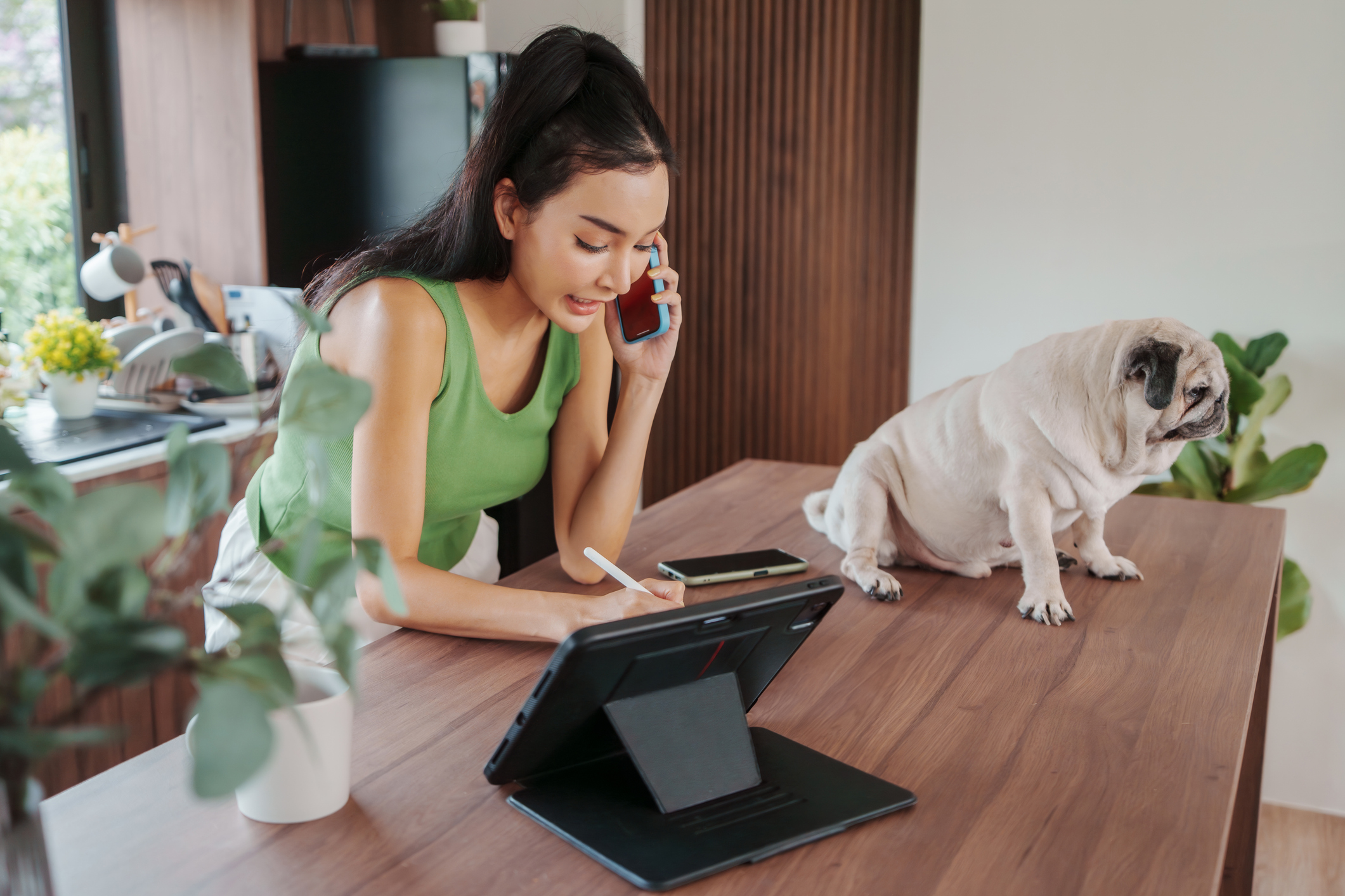 A young Asian woman engages in a pet care call while working on tablet with a pug at her side for proactive-outreach blog.
